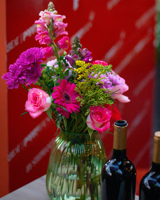 Bouquet of flowers in a green vase with wine bottles on a table against a red background.